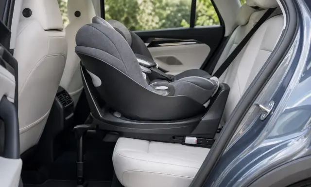 Silla de coche para bebé, instalada a favor de la marcha, con base giratoria y tapicería gris sobre asientos de cuero blanco en el interior de un vehículo.