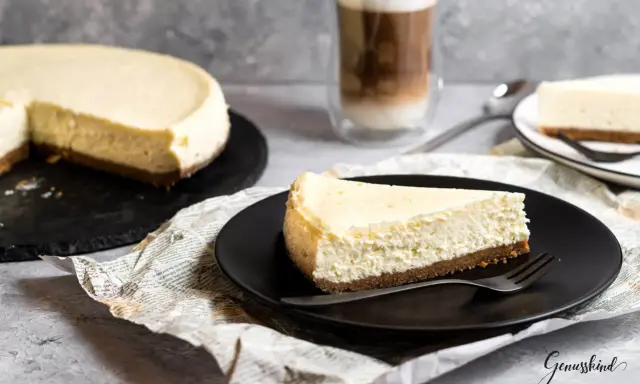 Ein Stück New York Cheesecake auf schwarzer Platte, daneben ein ganzer Kuchen und ein Latte Macchiato.