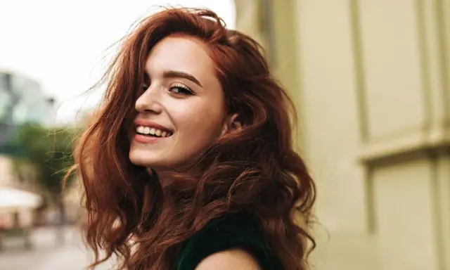 Donna con capelli rossi mossi e un sorriso radioso. La sua chioma è così bella che si può fare la tinta ogni 15 giorni per mantenerla perfetta.