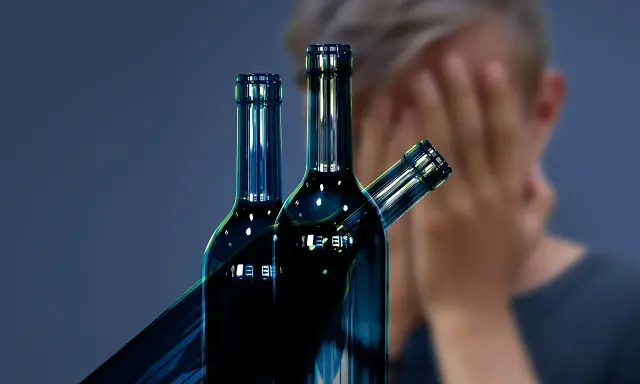 Trzy butelki alkoholu na pierwszym planie, w tle osoba zakrywająca twarz. Symbol walki z nałogiem, nadzieja na odwyk alkoholowy.