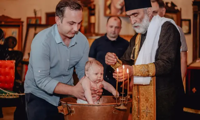 Chrzest dziecka w kościele. Ojciec trzyma niemowlę w miedzianej chrzcielnicy, a ksiądz polewa je wodą. To ważne chrzciny.