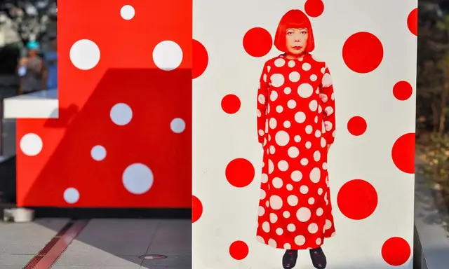 A woman in a red and white polka dot dress stands amidst Yayoi Kusama patterns.