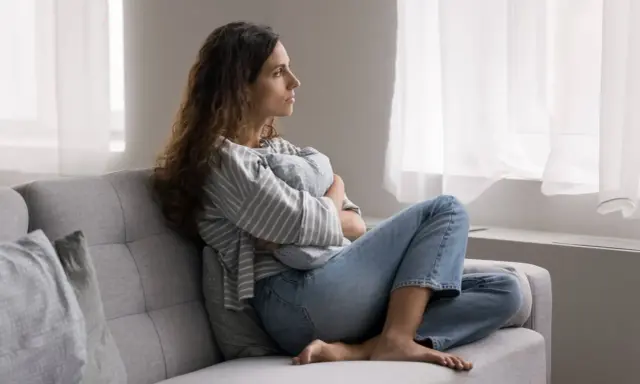 Kobieta siedzi na kanapie, przytulając poduszkę. Zamyślona patrzy przez okno. To pokazuje, po co światu psychoterapia – by odnaleźć spokój.