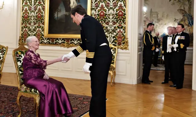 Drottning Margrethe, trots att hon är sjuk, tar emot en officer med ett leende. Hon sitter i en elegant stol, klädd i en lila klänning.