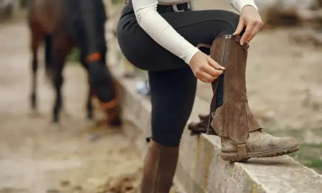 Jeździec zapina brązowe sztylpy na zamek, gotowy do jazdy. W tle widać konia.