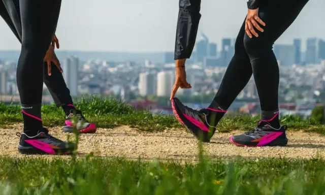 Zwei Läuferinnen dehnen sich auf einem Feldweg mit Blick auf eine Stadt. Sie tragen schwarze Laufhosen und auffällige pink-schwarze Laufschuhe. Ein Zeichen für den Wiedereinstieg ins joggen nach Geburt.