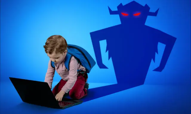 Chłopiec z plecakiem pochyla się nad laptopem, a za nim czai się złowrogi cień potwora. Zagrożenia czyhające na dzieci w internecie.