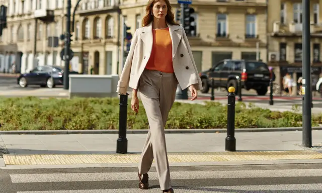 Elegancka kobieta w beżowych stylizacjach zimowych przechodzi przez pasy. Jasny płaszcz narzucony na pomarańczową bluzkę i spodnie w prążki tworzą szykowny miejski look.