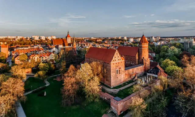 Zachód słońca nad zamkami krzyżackimi na Mazurach. Potężne ceglane mury i wieże w blasku złota, otoczone zielenią i miastem.