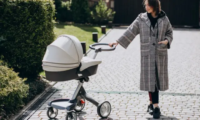 Kobieta w płaszczu prowadzi spacerówkę kubelkową po brukowanej ścieżce wśród zieleni.