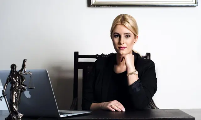 Kobieta w czarnej marynarce siedzi przy biurku z laptopem i figurką Temidy.