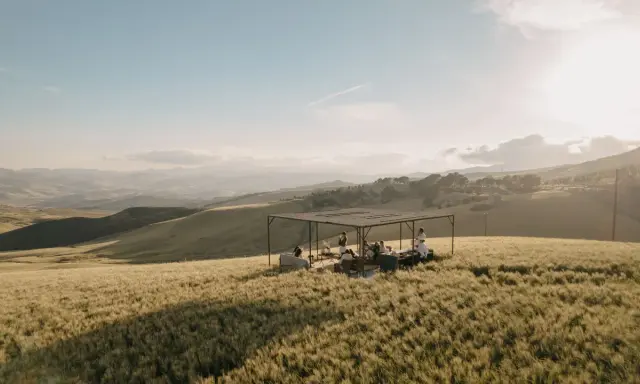 Un gruppo di persone si riunisce sotto una tettoia in un campo di grano dorato, godendosi la tipica bellezza siciliana al tramonto.