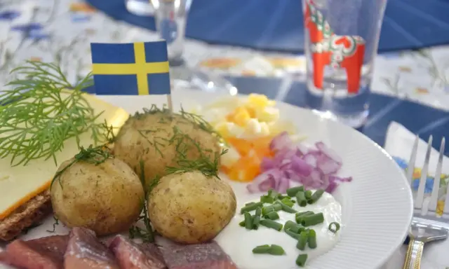En tallrik med midsommar mat: sill, färskpotatis, ost, ägg och rödlök. En svensk flagga vajar stolt.