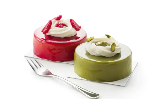 Deux petits gâteaux brillants, l'un rouge vif, l'autre vert avec un glaçage pistache, ornés de crème et de fruits.