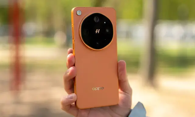Pomarańczowy oppo flagowiec z charakterystycznym modułem aparatu w dłoni, na tle rozmytego, zielonego krajobrazu.