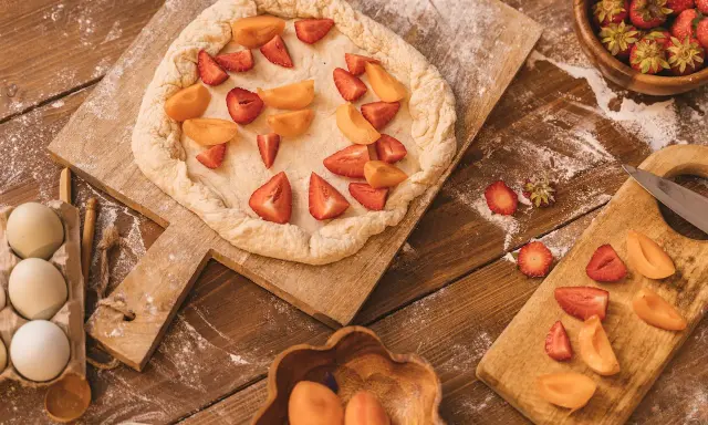 Préparation d'un crumble abricot et fraises sur une planche en bois, avec des œufs et des fruits frais à proximité.