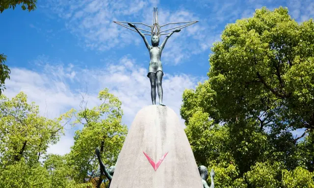 La bambina di Hiroshima, simbolo di pace, con le braccia tese verso il cielo, circondata dal verde degli alberi e dall'azzurro delle nuvole.
