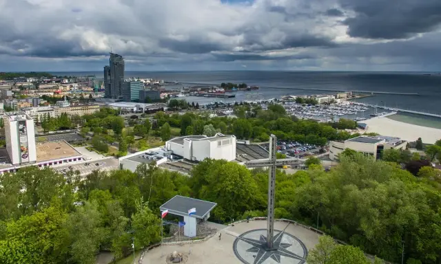 Gdynia co zobaczyć: pomnik z różą wiatrów i krzyżem na wzgórzu, w tle port, plaża i drapacze chmur.