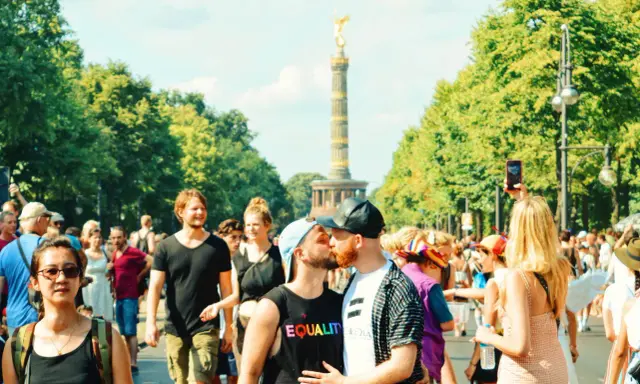 A couple kisses at a pride parade in Germany, with the Victory Column in the background. This scene shows Germany is gay friendly.