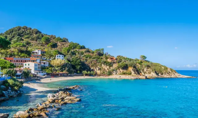 En pittoresk kustby på Elba, Italien, med vita hus som klättrar uppför en grönskande kulle. Klart turkost vatten sköljer mot en sandstrand.
