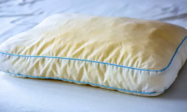 Ein vergilbtes Kissen mit blauen Nähten, das auf einem weißen Bett liegt. Gelbe Flecken deuten auf möglichen Schimmel hin.