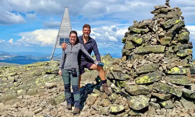 Två glada vandrare njuter av utsikten från en bergstopp under en vandring i Sverige. En stenröse och en modern träkonstruktion med solpaneler syns i bakgrunden.