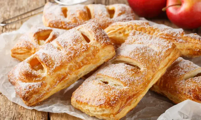 Chrupiące ciasto francuskie z jabłkami, posypane cukrem pudrem, czeka na spróbowanie. Obok leżą rumiane jabłka.