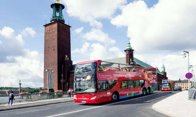 Röd dubbeldäckare med "Hop-on Hop-off Stockholm Buses" på väg förbi Stadshuset. Perfekt för stockholm turistinformation.