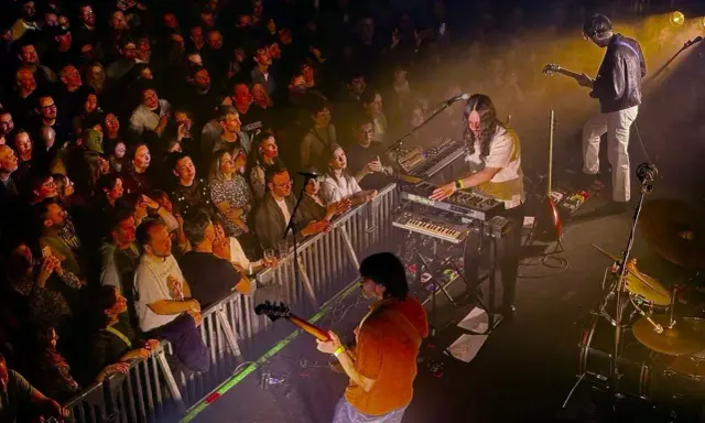 Publik njuter av Altin Gün i Stockholm. En gitarrist spelar, en keyboardist bakom Roland-syntar, och en entusiastisk publik bakom stängsel.