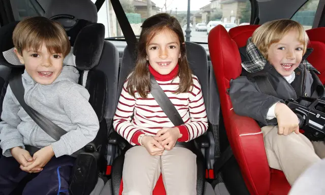 Tres niños sonríen en sus asientos de coche, listos para viajes en coche con niños de 2 años.