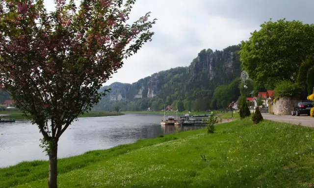 Kwiaty na drzewie i zielona trawa nad rzeką, w tle skaliste góry i miasteczko. Idealne miejsce na spacer, jak z **bastei cennik** wycieczki.