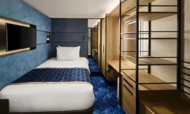 Interno cabina singola nave da crociera: letto, TV a parete, armadio aperto con mensole e scrivania. Le dimensioni delle cabine sono ottimizzate per il comfort.