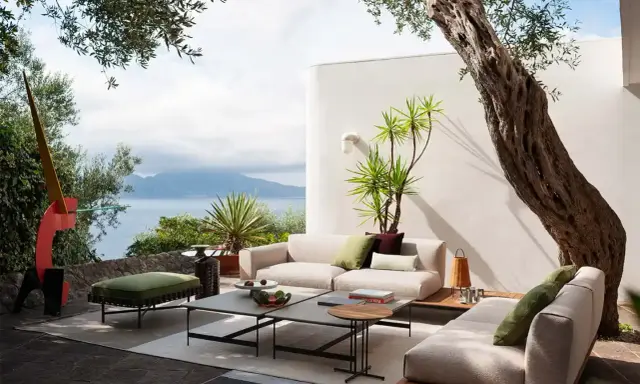 Arredare un terrazzo scoperto con divani chiari, tavolini bassi e un pouf verde, con vista mare e un ulivo secolare.