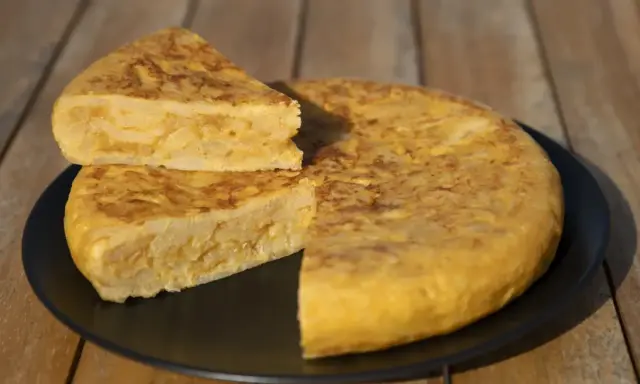 Una deliciosa tortilla de patatas, con una porción cortada y apilada, servida en un plato negro sobre una mesa de madera.