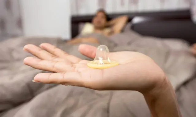 Mano sostiene un condón. En el fondo, una persona en la cama. La decisión de tener sexo oral sin condón es personal.