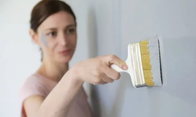 Une femme effectue une retouche peinture murale avec un pinceau gris. Une tache de peinture grise est visible sur sa joue.