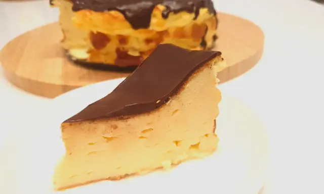 Kawałek puszystego keto sernika z mascarpone, polany czekoladą. Cały sernik w tle.