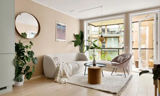 Jasne meble do salonu: puszysta sofa, okrągły stolik, aksamitny fotel i puf. Duże okno z widokiem na balkon.
