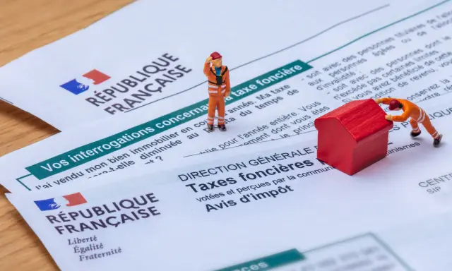 Figurines sur des documents de la République Française concernant la taxe foncière. L'une pousse une maison rouge, l'autre semble réfléchir.
