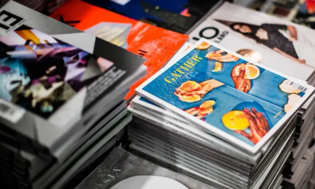 Stosy magazynów, jeden z nich z okładką "GATHER" i pomarańczami, gdzie każdy detal, od tekstury owoców po odbicia światła, podkreśla piękno koloru spotowego.