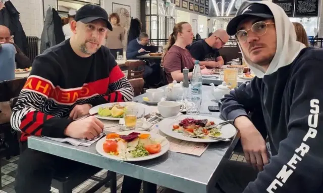 Dwóch mężczyzn, jeden w bluzie z napisem "Givenchy", drugi w bluzie "Supreme", siedzi przy stole w restauracji. Wyglądają jak bary raperzy, którzy wybrali się na wspólny posiłek.