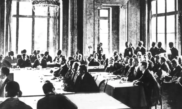Männer in Anzügen sitzen an langen Tischen in einem prunkvollen Saal. Dies ist die Unterzeichnung des Versailler Vertrags, der den Ersten Weltkrieg beendete.