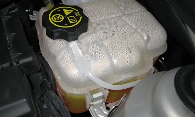Depósito de expansión con líquido refrigerante y burbujas. La tapa negra tiene símbolos de advertencia.