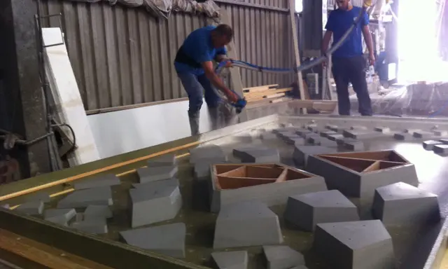 Dos operarios trabajan con material GRC en un taller, uno rocía con una manguera y el otro supervisa.