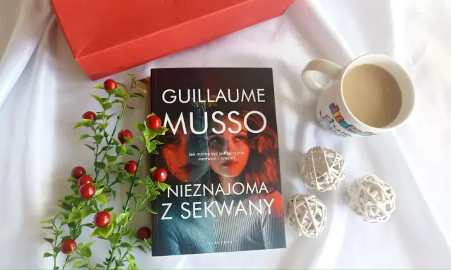 Książka "Nieznajoma z Sekwany" Guillaume'a Musso leży obok kubka kawy i czerwonych ozdobnych kulek.