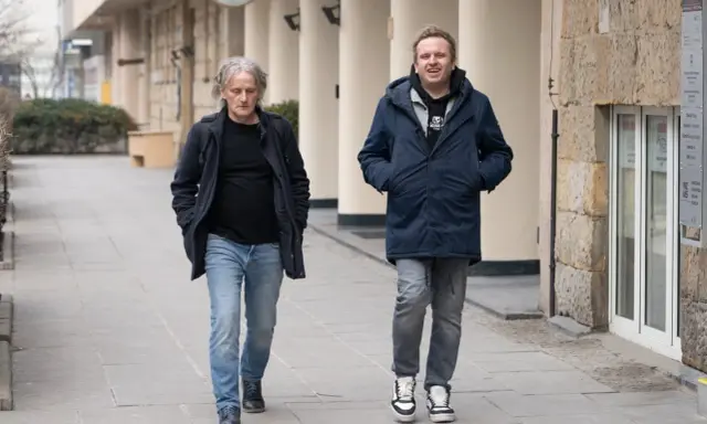 Jacek Podsiadło, ojciec Dawida, idzie ulicą w granatowej kurtce i jeansach. Obok niego młodszy mężczyzna w podobnym stroju.