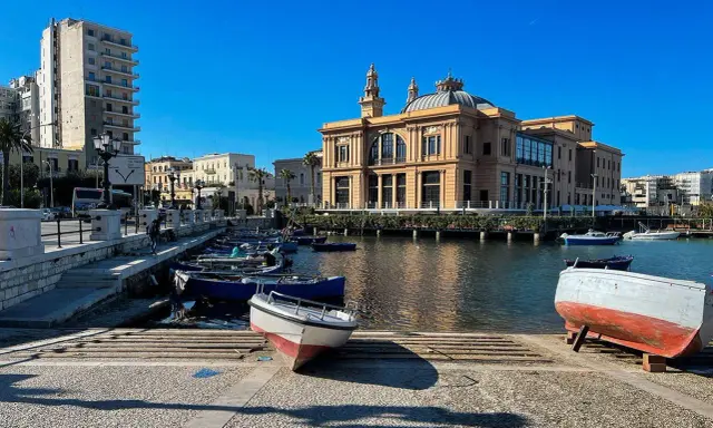 Barche ormeggiate nel porto di Bari, con il Varco della Vittoria e il Teatro Margherita sullo sfondo, sotto un cielo azzurro.