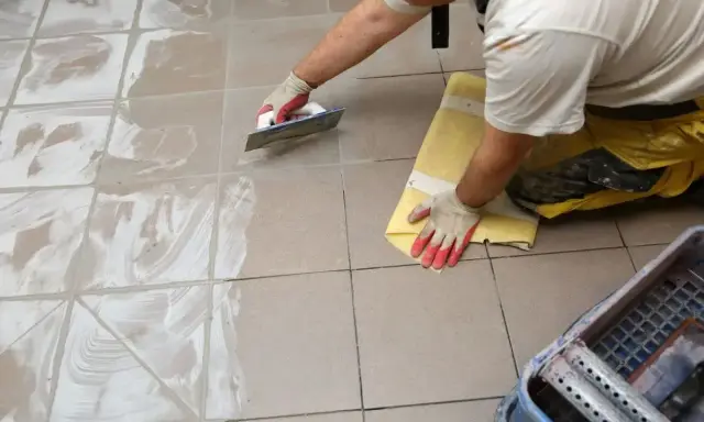 Pracownik w rękawiczkach nakłada fugę epoksydową na płytki podłogowe za pomocą kielni. Proces fugowania krok po kroku.