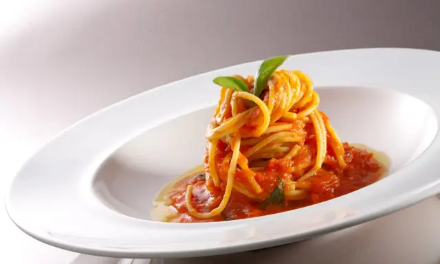 Un nido di spaghetti conditi con una salsa rossa vibrante di pomodorini freschi, guarnito con foglie di basilico verde brillante, servito in un piatto bianco elegante.