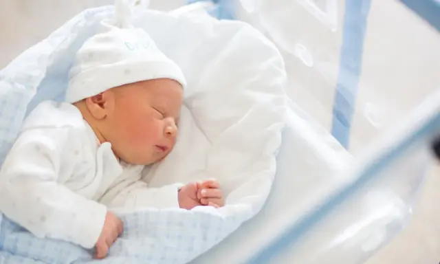 Śpiący noworodek w szpitalnym łóżeczku. Do wniosku o becikowe potrzebne są dokumenty potwierdzające urodzenie dziecka.
